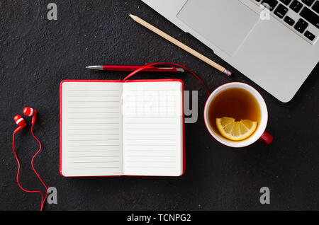 Leeres rotes Notizbuch mit Stift, Computer, Laptop, Kopfhörer und Tasse Tee auf dunklen Tisch. Geschäft noch leben, Büro oder Bildung Konzept. Blick von oben auf die w Stockfoto