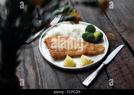 Kabeljau paniert gebraten mit Reis, Brokkoli und frische Zitrone auf einem weißen Teller serviert. Horizontale, Holz- Hintergrund Stockfoto