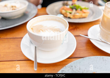 Fokus im Vordergrund auf Kaffee Cappuccino. Eine junge Frau mit Frühstück in einem Sommer Cafe. Stockfoto