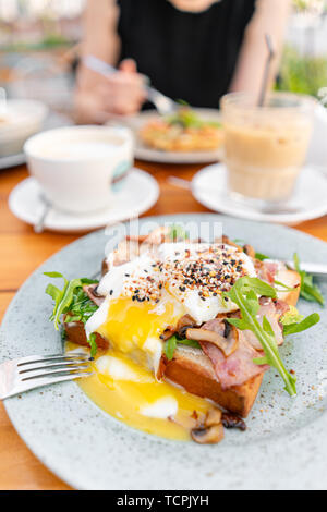 Frühstück und Kaffee im Sommer Cafe. Sandwich in einem Brötchen Brioche mit Speck, Champignons, Rucola und pochiertem Ei Stockfoto