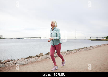 Gesunde blond zeitgenössische Sportlerin Jogging entlang Riverside beim Hören von Musik über Kopfhörer während des Trainings Stockfoto