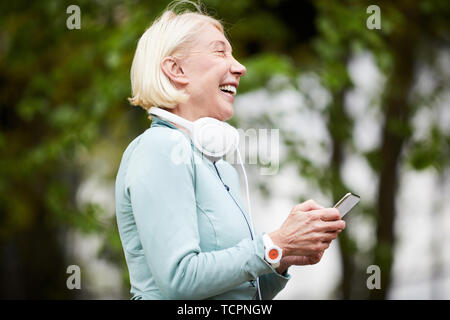 Reifen ekstatisch Sportlerin mit Smartphone Lachen beim Scrollen oder SMS in der natürlichen Umgebung im Sommer Tag Stockfoto