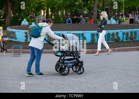 Nowosibirsk, Russland - 05.28.2019: Junge Mutter Mädchen mit Kinderwagen und Kleinkindern sind zu Fuß in einer Stadt Park vor dem hintergrund der grünen Bäumen, Stockfoto