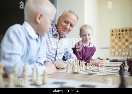 Lächelnd Großvater in kariertes Hemd Lehre seine zwei neugierige grandkids, wie man Schach spielen Stockfoto