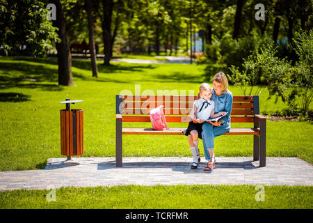 Mutter und Tochter im Freien öffentlichen Park auf der Bank und lesen Buch sitzen, Lektionen. Elternschaft und Kind Konzept. Stockfoto