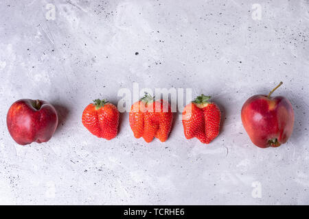 Hässliche zwei Äpfel und drei Erdbeeren liegen in Reihe auf grauem Beton Hintergrund. Stockfoto