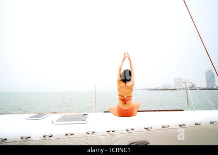 Rückseite des jungen, gesunden und ruhigen frau yoga meditation und Sitzen mit den Händen auf Segelyacht Boot mit Ozean Meer im Hintergrund Stockfoto