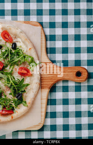 Neapolitanische Pizza auf Sahnesoße mit Thunfisch, Käse, Rucola, Basilikum, Tomaten, Oliven, bestreut mit Käse auf einem Holztisch auf einer Tischdecke in einer Zelle Stockfoto