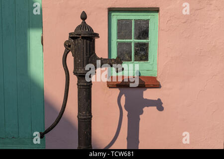 Eine altmodische Wasserpumpe gegen eine rosa Wand mit einem grünen Fenster, mit einem starken Schatten der Pumpe an der Wand, niemand im Bild Stockfoto