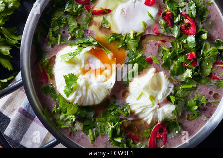 Frische hausgemachte Mexikanische scharfe rote Kidney-bohnen-Suppe mit pochierten Eiern und Koriander in eine Metallwanne auf einer konkreten Tabelle mit einem Holzlöffel und Inhaltsstoffe Stockfoto