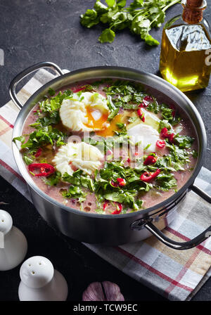 Hot mexikanische scharfe rote Kidney-bohnen-Suppe mit pochierten Eiern und Koriander in eine Kasserolle auf einer konkreten Tabelle mit einem Holzlöffel und Zutaten, ve Stockfoto
