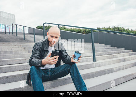 Nach kahlen düsteren Mann trinken Kaffee aus dem Pappbecher und über Handy sitzen in Treppen an den Stadt. Stockfoto