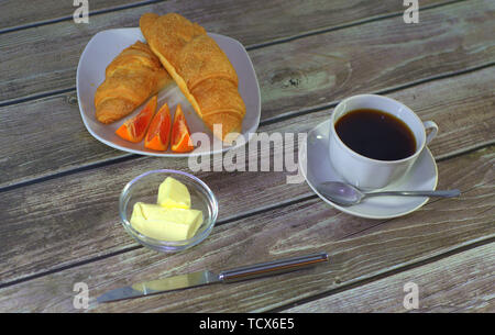 Eine Tasse schwarzen Kaffee auf einer Untertasse mit einem Löffel, eine Platte mit Croissants und Zitronenscheiben mit Butter stehen auf einem Holztisch. Close-up. Stockfoto