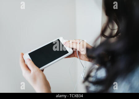 Junge Frau mit weißen Smartphone, auf den Bildschirm zu schauen. Weißer Hintergrund, kein Gesicht gesehen, Kopfhörer auf ihren Arm. Stockfoto