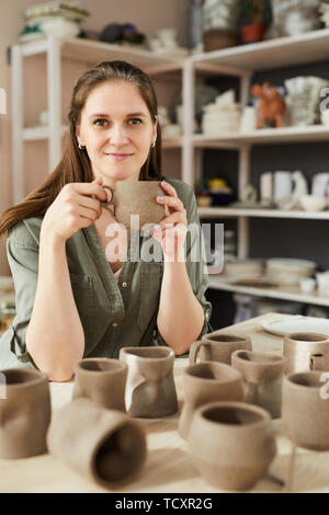 Portrait von lächelnd weibliche Handwerker Holding handgefertigte Keramik Becher, während im traditionellen Töpferei shop Posing Stockfoto