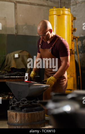 Konzentriert kahlen Handwerk Arbeiter in der Schürze am Amboss und Ändern der Form von erhitztem Metall mittels Hammer Stockfoto