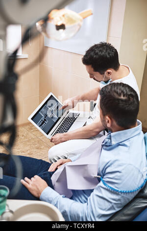 Dunkle bärtigen Haaren Zahnarzthelfer tragen weiße Uniform bereit ist, die Zähne des Patienten zu überprüfen. Er zeigt X-ray Bilder der Zähne seiner Patienten mit La Stockfoto