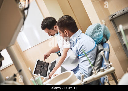Dunkle bärtigen Haaren Zahnarzthelfer tragen weiße Uniform bereit ist, die Zähne des Patienten zu überprüfen. Er zeigt X-ray Bilder der Zähne seiner Patienten mit La Stockfoto