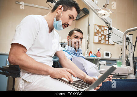 Dunkle bärtigen Haaren Zahnarzthelfer tragen weiße Uniform bereit ist, die Zähne des Patienten zu überprüfen. Er zeigt X-ray Bilder der Zähne seiner Patienten mit La Stockfoto