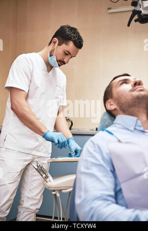 Dunkle bärtigen Haaren Zahnarzthelfer tragen weiße Uniform bereit ist, die Zähne des Patienten zu überprüfen. Stockfoto