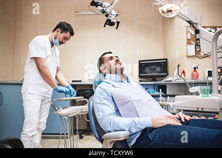 Dunkle bärtigen Haaren Zahnarzthelfer tragen weiße Uniform bereit ist, die Zähne des Patienten zu überprüfen. Stockfoto