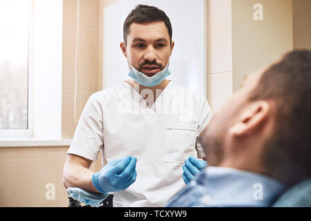 Dunkle bärtigen Haaren Zahnarzthelfer tragen weiße Uniform bereit ist, die Zähne des Patienten zu überprüfen. Er sitzt in der Nähe von zahnmedizinischen Stuhl im Gespräch mit seinem Patienten ein Stockfoto
