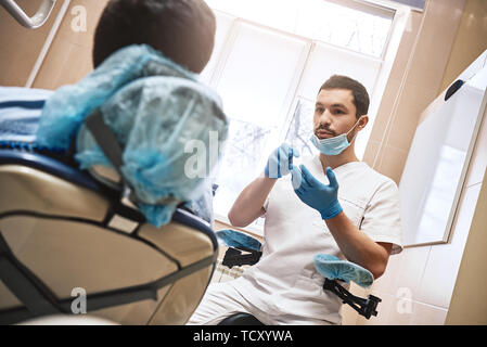 Dunkle bärtigen Haaren Zahnarzthelfer tragen weiße Uniform bereit ist, die Zähne des Patienten zu überprüfen. Er sitzt in der Nähe von zahnmedizinischen Stuhl im Gespräch mit seinem Patienten ein Stockfoto