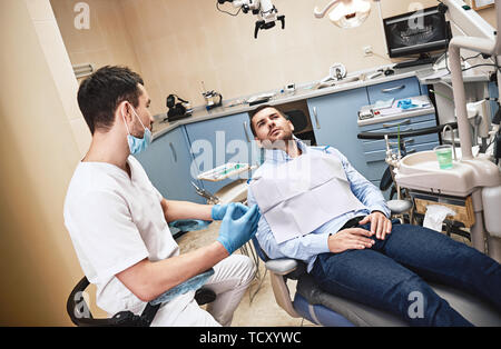 Dunkle bärtigen Haaren Zahnarzthelfer tragen weiße Uniform bereit ist, die Zähne des Patienten zu überprüfen. Er sitzt in der Nähe von zahnmedizinischen Stuhl im Gespräch mit seinem Patienten ein Stockfoto
