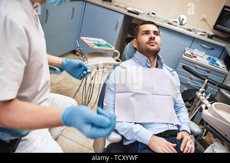Dunkle bärtigen Haaren Zahnarzthelfer tragen weiße Uniform bereit ist, die Zähne des Patienten zu überprüfen. Er sitzt in der Nähe von zahnmedizinischen Stuhl im Gespräch mit seinem Patienten ein Stockfoto