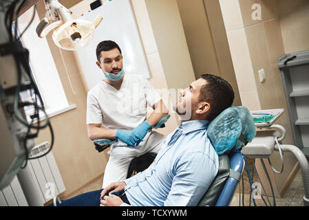 Dunkle bärtigen Haaren Zahnarzthelfer tragen weiße Uniform bereit ist, die Zähne des Patienten zu überprüfen. Er sitzt in der Nähe von zahnmedizinischen Stuhl Gespräch mit seinem Patienten. Sid Stockfoto