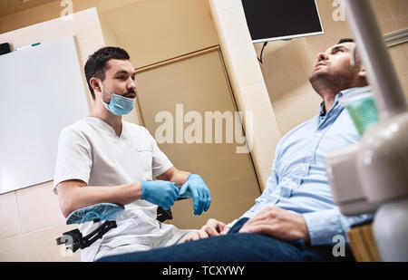Dunkle bärtigen Haaren Zahnarzthelfer tragen weiße Uniform bereit ist, die Zähne des Patienten zu überprüfen. Er sitzt in der Nähe von zahnmedizinischen Stuhl Gespräch mit seinem Patienten. Sid Stockfoto