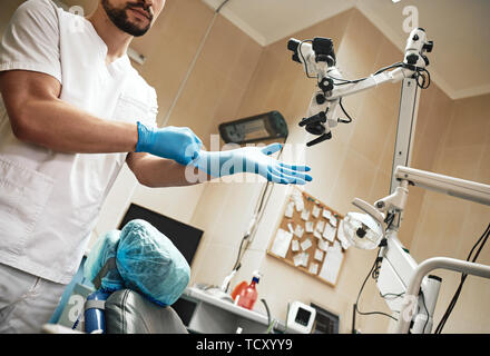 Bärtige Zahnarzthelfer tragen weiße Uniform ist bereit, bis die Zähne des Patienten zu überprüfen. Er trägt OP-Handschuhe während der Wartezeit für die Patienten. Nahaufnahme Stockfoto