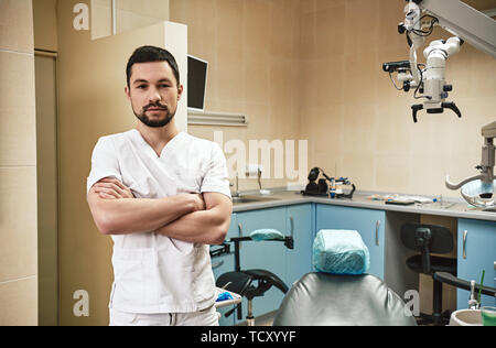 Dunkle bärtigen Haaren Zahnarzthelfer tragen weiße Uniform bereit ist, die Zähne des Patienten zu überprüfen. Er steht mit verschränkten Armen, während für Pat warten Stockfoto