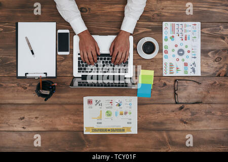 Blick von oben auf die menschliche Hände schreiben auf Laptop. Laptops, Smartphones, Tagebuch, Tasse, Gläser, Charts und andere Verbrauchsmaterialien auf braun Holzschreibtisch. Mann bei der Arbeit Stockfoto