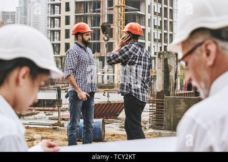 Anstrengenden Tag. Team von professionellen Ingenieuren und Bauherren in Schutzkleidung arbeiten gemeinsam an der Baustelle. Junge builder spricht per Telefon. Stockfoto