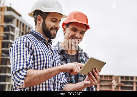 Arbeiten im Team. Zwei junge und fröhliche Bauherren in schützende Helme sind mit digitalen Tablet und Arbeiten an der Baustelle stehen. Konst Stockfoto