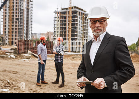 Besuch der Baustelle. Portrait von zuversichtlich ältere Bauingenieur im klassischen Anzug und hardhat hält digital Tablet und c Stockfoto