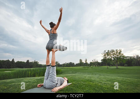 Schönen Posen mit einem Partner. Starke Mann liegt auf einer Matte und Balancing Frau in Lotus auf die Beine stellen. Junges Paar, acro Yoga in der Natur. Gesundheit Stockfoto