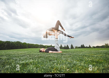 Fit Paar. Starken jungen Mann liegen auf Gras und Balancing Frau auf seine Beine. Gesundes Paar üben acro Yoga in der Natur. Gesunder Lebensstil Konzept. Stockfoto