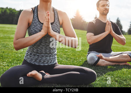 Yoga im Park. Nahaufnahme des jungen Paares üben Meditation Übungen im Freien sitzen Sitzen im Lotussitz auf grünem Rasen im freien Feld. Med Stockfoto