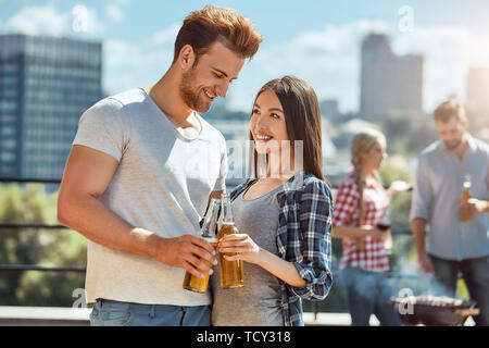 Cheers zu uns! Junge und schöne Paar klirren Gläser mit Bier und lächelnd, während auf dem Dach mit Freunden. Grill Konzept. Das Kochen. P Stockfoto