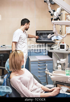 Dunkle bärtigen Haaren Zahnarzthelfer tragen weiße Uniform bereit ist, die Zähne des Patienten zu überprüfen. Er zeigt X-ray Bilder der Zähne seiner Patienten mit Co Stockfoto