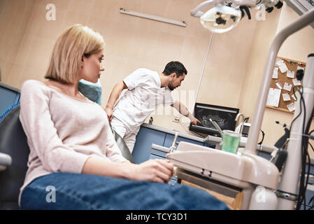 Dunkle bärtigen Haaren Zahnarzthelfer tragen weiße Uniform bereit ist, die Zähne des Patienten zu überprüfen. Er zeigt X-ray Bilder der Zähne seiner Patienten mit Co Stockfoto