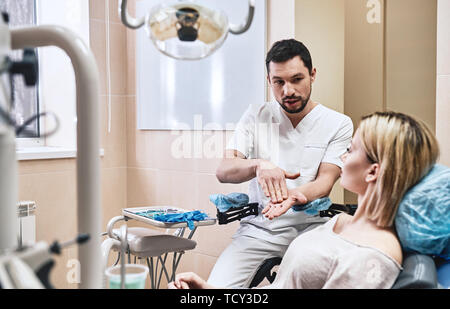 Dunkle bärtigen Haaren Zahnarzthelfer tragen weiße Uniform bereit ist, die Zähne des Patienten zu überprüfen. Er sitzt in der Nähe von zahnmedizinischen Stuhl im Gespräch mit seinem Patienten ein Stockfoto