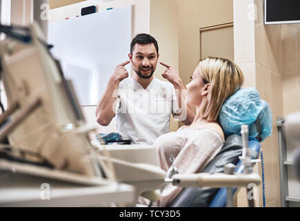 Dunkle bärtigen Haaren Zahnarzthelfer tragen weiße Uniform bereit ist, die Zähne des Patienten zu überprüfen. Er sitzt in der Nähe von zahnmedizinischen Stuhl im Gespräch mit seinem Patienten ein Stockfoto