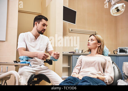 Dunkle bärtigen Haaren Zahnarzthelfer tragen weiße Uniform bereit ist, die Zähne des Patienten zu überprüfen. Er sitzt in der Nähe von zahnmedizinischen Stuhl im Gespräch mit seinem Patienten ein Stockfoto