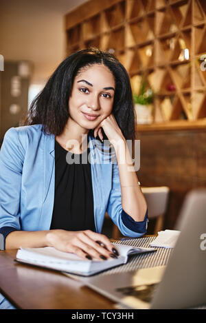 Porträt der jungen Brünett trägt blaue Jacke, in die Kamera schaut. Sie ist Notizen zu machen, mit Laptop Computer während der Mittagspause. Vertikaler Dampfstoß Stockfoto