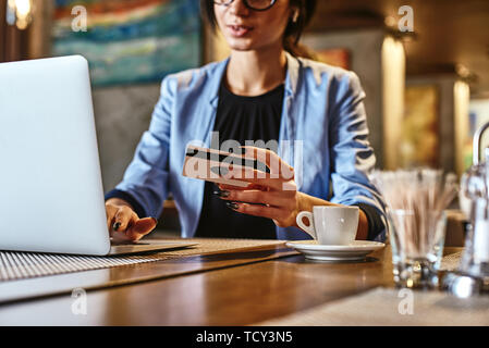 Zugeschnittenes Bild der weiblichen Holding Kreditkarte, die Transaktion, über Mobile Banking App beim Mittagessen im Cafe. Ihr Kaffee und Laptop auf dem Tisch Stockfoto
