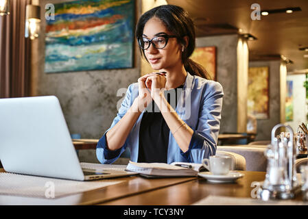 Positive stilvolle Geschäftsfrau in blaue Jacke und Brille sitzen am Tisch, im Innenbereich, trägt drahtlose Kopfhörer Chat mit Ihrem Chef über Video c Stockfoto