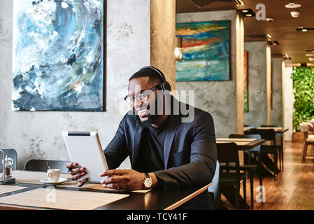Portrait von positiven Stilvolle Junge Afro-amerikanische Geschäftsmann in dunkel-blaue Jacke und Brille sitzen am Tisch, drinnen, das Tragen von kabellosen Ohrhörern, Stockfoto